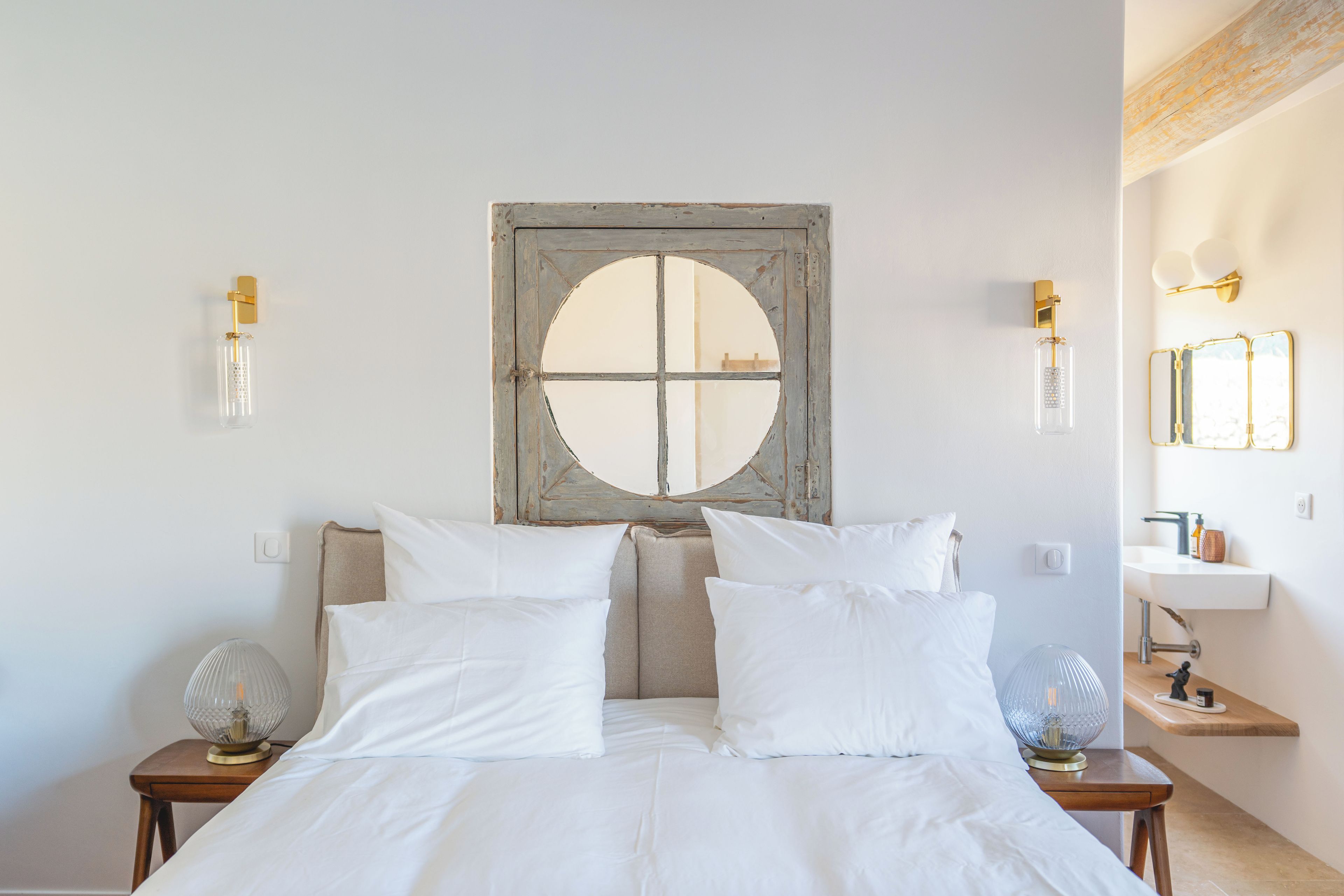 Une chambre à coucher avec un grand lit, deux tables de chevet, un miroir fixé au mur et une fenêtre avec un cadre en bois.