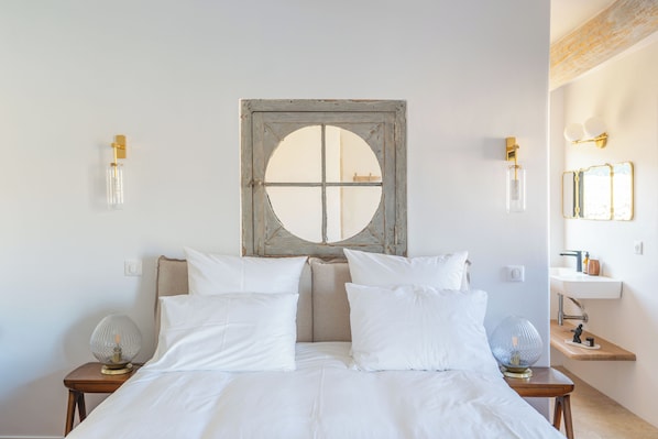 A bedroom with a large bed, two bedside tables, a wall-mounted mirror, and a window with a wooden frame.