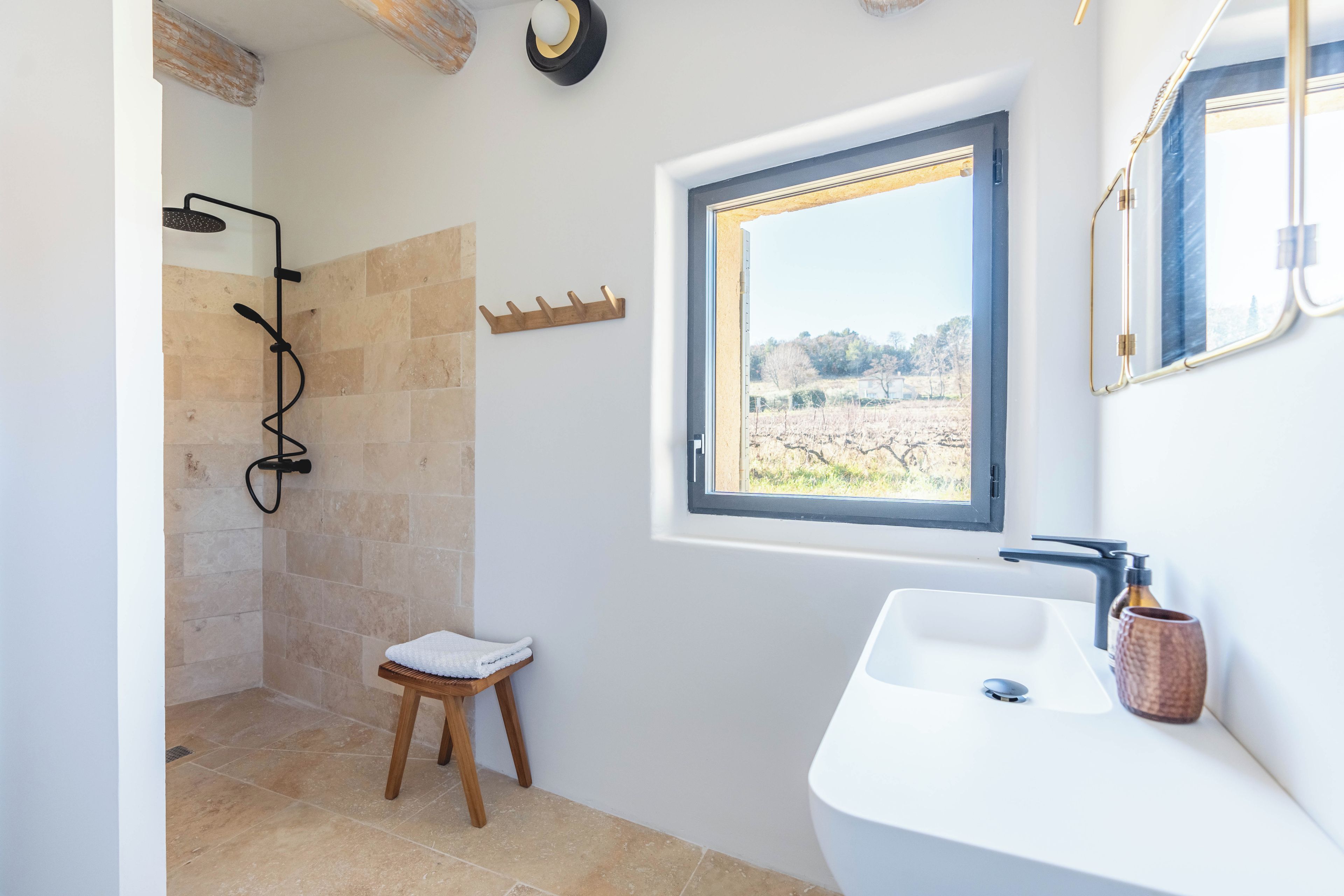 A bathroom with a shower, a small stool, a sink, and a window with a view of the outdoors.