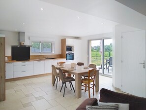 Eine moderne Küche mit weißen Schränken, ein Essbereich mit Holztisch und -stühlen sowie eine Glastür, die auf eine Terrasse im Freien führt.