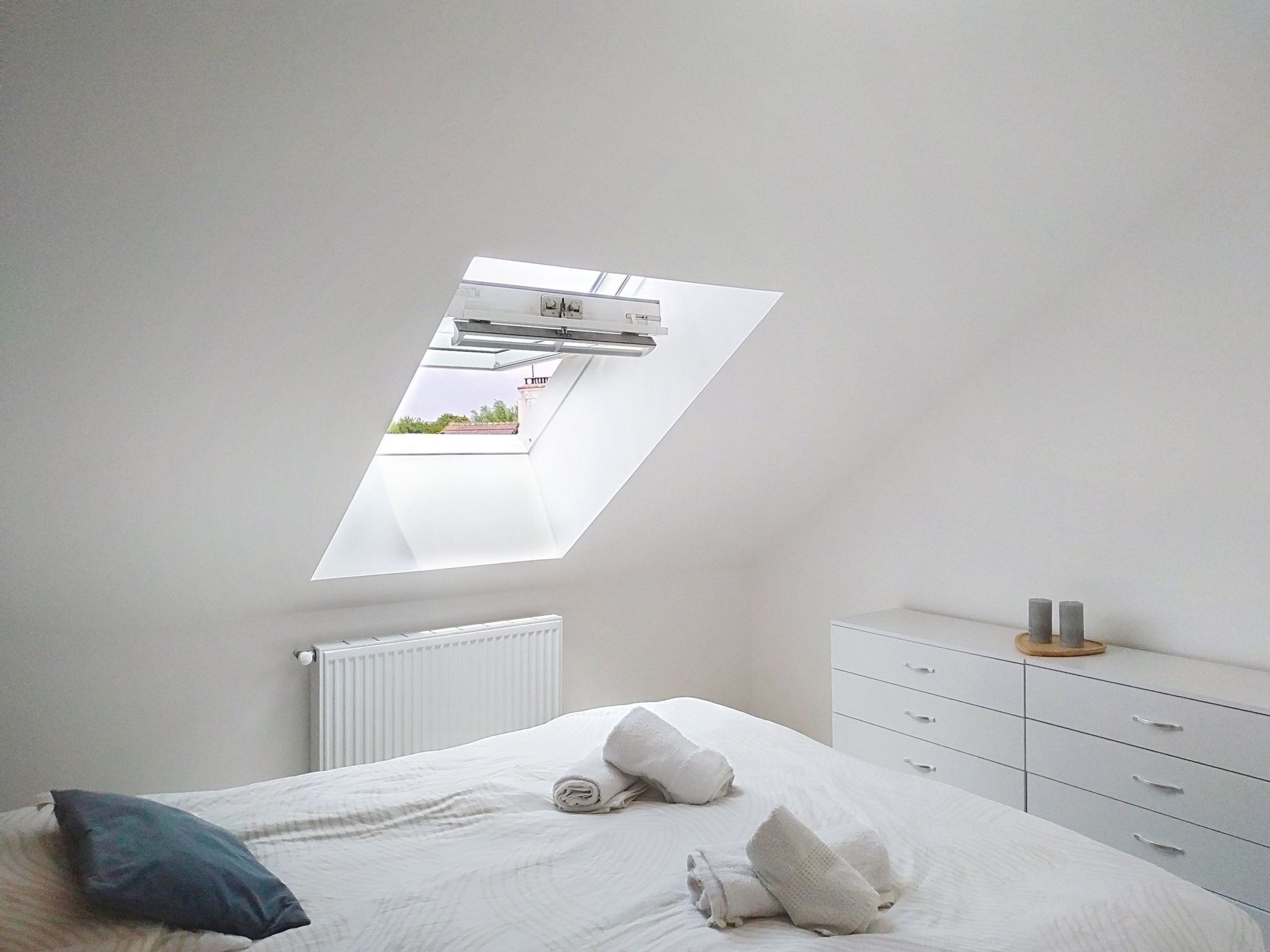 Quarto com claraboia, radiador, cama com almofadas e cómoda com velas.