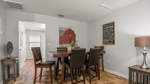 Un coin repas avec une table et des chaises en bois, un tableau représentant une grange rouge, un panneau noir et un lampadaire sur une table d’appoint.