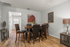 Ein Essbereich mit Holztisch und -stühlen, einem Gemälde von einem roten Bauernhaus, einem Kreidetafel-Schild und einer Lampe auf einem Beistelltisch.