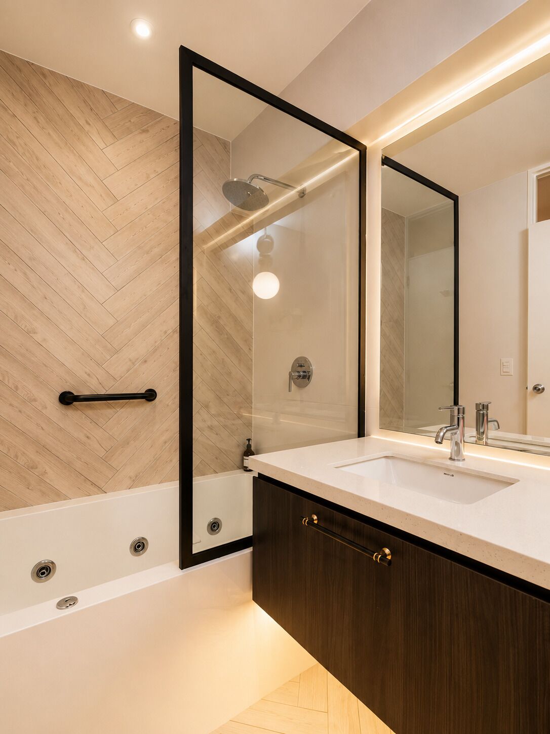 Ein modernes Badezimmer mit Fischgrätparkett, einer Glasdusche, einem weißen Waschbecken mit dunklem Holzschrank und einem großen Spiegel.