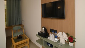 Ein Hotelzimmer mit Flachbildfernseher an einer Holzverkleidung, einem kleinen Tisch mit Wasserkocher und Fernbedienung, einem Sessel und einem Fenster mit Vorhängen.
