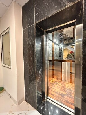 An elevator with a glass door, a wooden floor inside, and a reflection of a room with a red object. - SHREE LEELAA HOTEL (Jaipur)