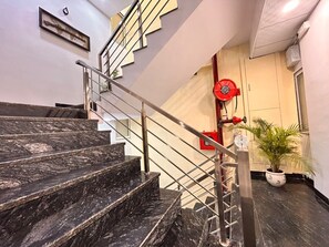 A staircase with marble steps and metal railings, leading to a landing with a red fire alarm and a potted plant. - SHREE LEELAA HOTEL (Jaipur)