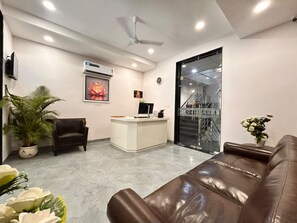 A reception area with a reception desk, a seating area with a couch, a potted plant, and a staircase visible through a glass door. - SHREE LEELAA HOTEL (Jaipur)