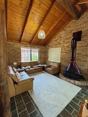 A cozy living room with a sectional sofa, a fireplace, and a view of the outdoors through a window. - Gumnut Cottage - 2BR Cottage, walk to beach and town, Linen & Wifi incl (Dunsborough)