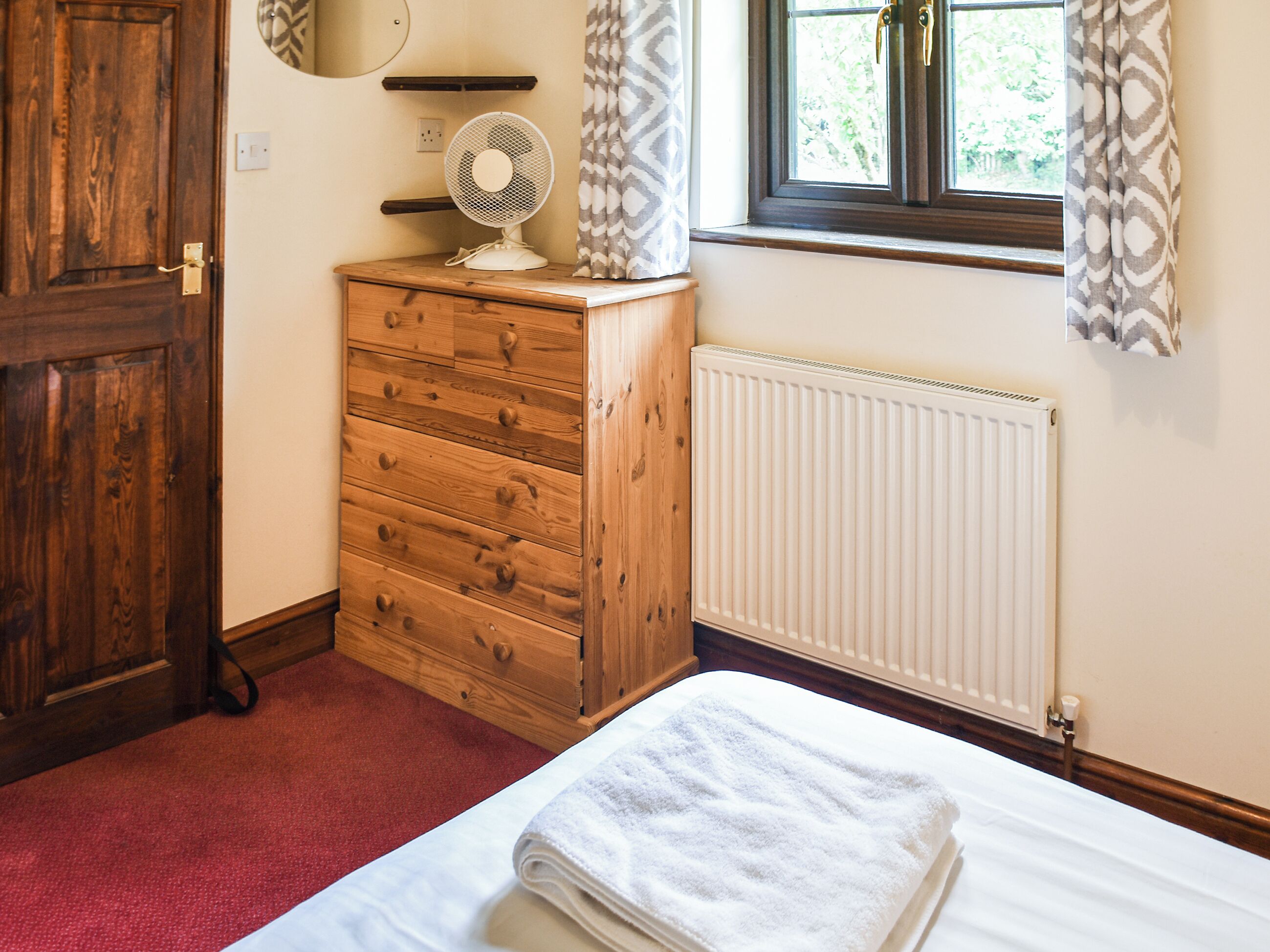 A bedroom with a wooden dresser, a radiator, a window with curtains, and a bed with a folded towel.
