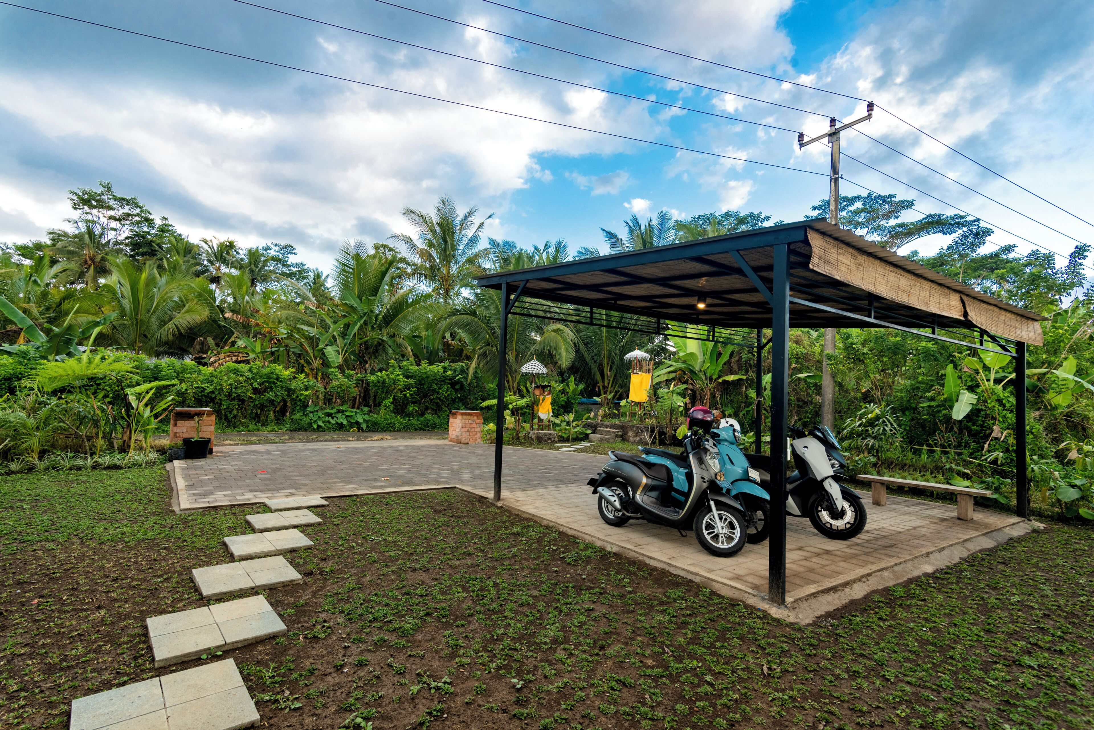 Un espacio de estar al aire libre techado, con una moto estacionada debajo, rodeado de abundante vegetación y un sendero pavimentado.