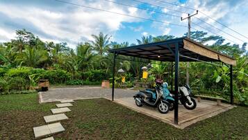 Un espacio de estar al aire libre techado, con una moto estacionada debajo, rodeado de abundante vegetación y un sendero pavimentado.