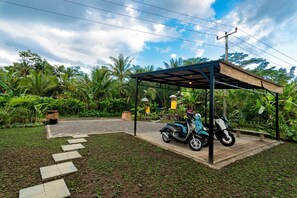 Un espacio de estar al aire libre techado, con una moto estacionada debajo, rodeado de abundante vegetación y un sendero pavimentado.
