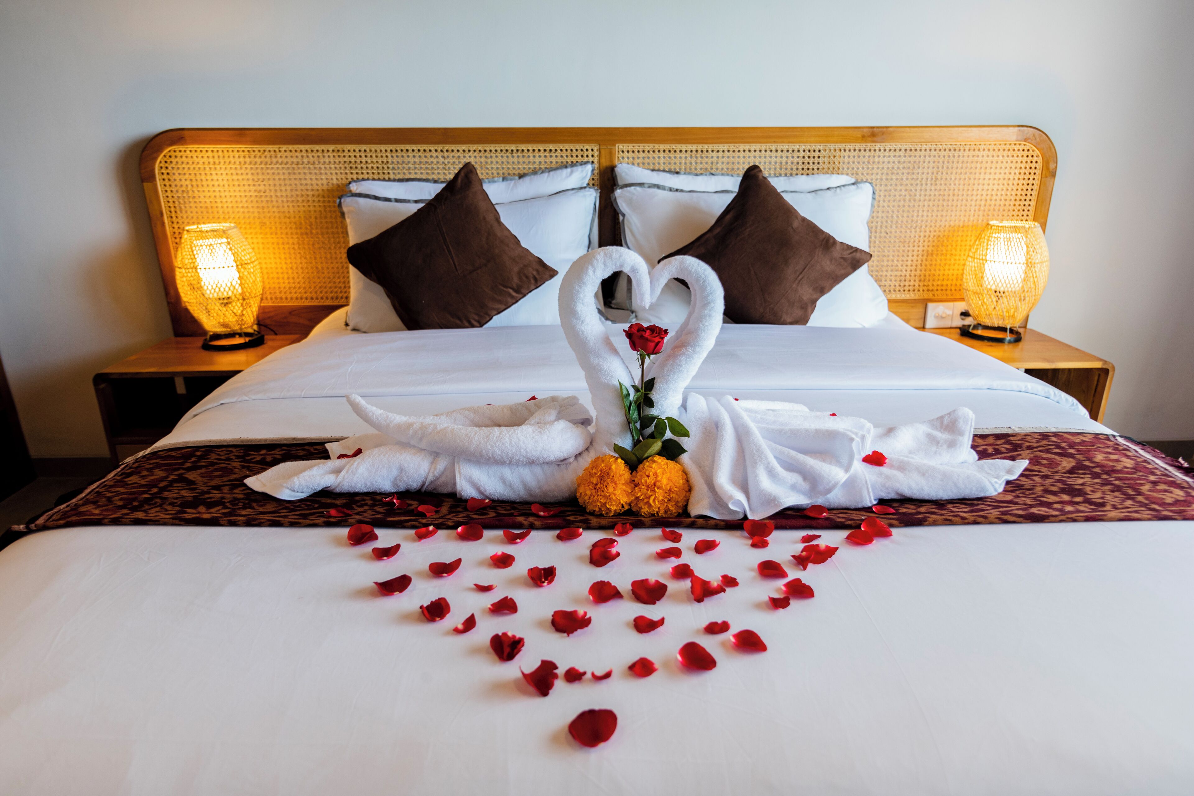 Una cama impecable con un arreglo de toallas en forma de corazón y pétalos de rosa, dos almohadas marrones y dos lámparas de noche.
