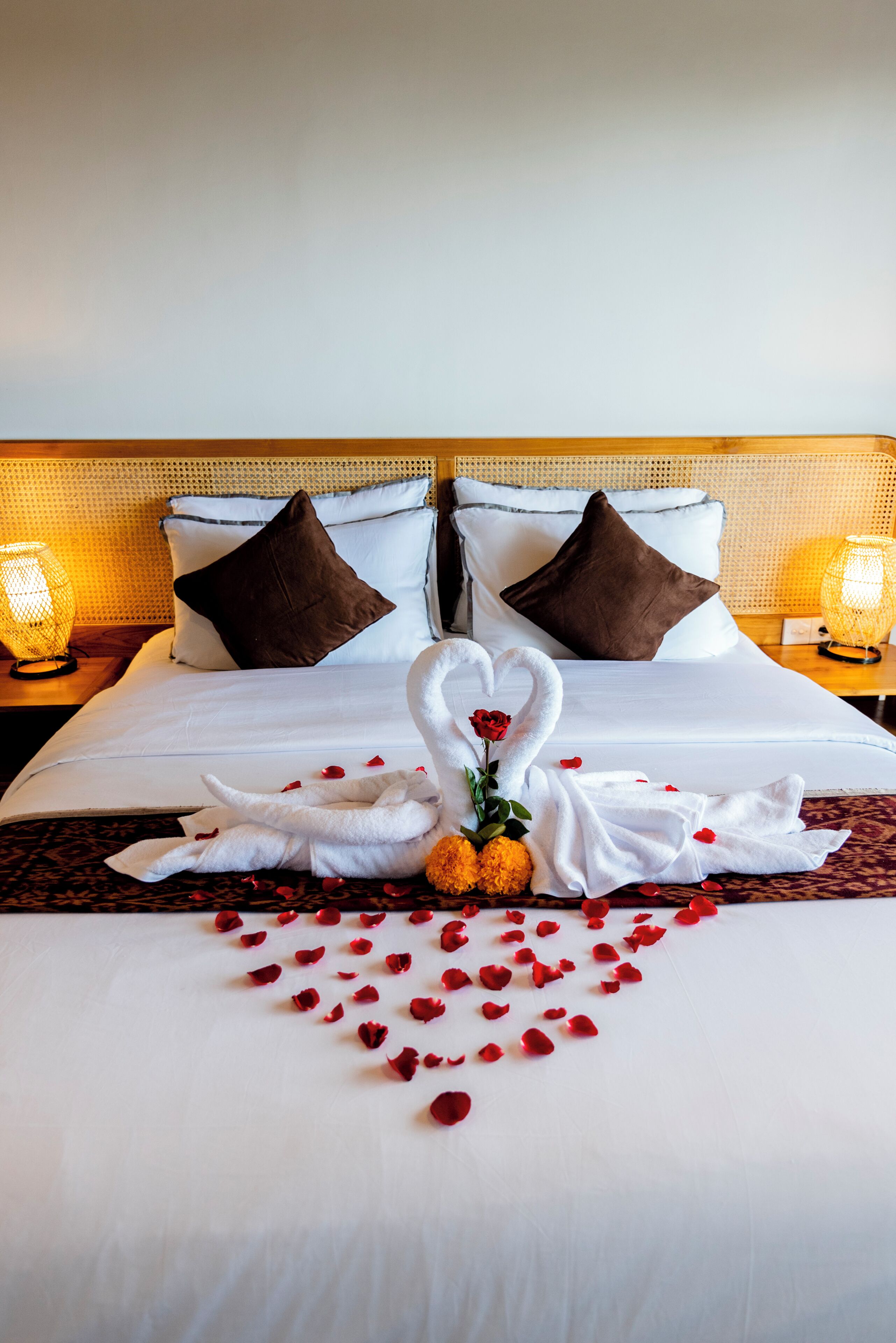Una cama impecable con una decoración de toallas en forma de corazón, pétalos de rosa y un centro de mesa con una rosa.