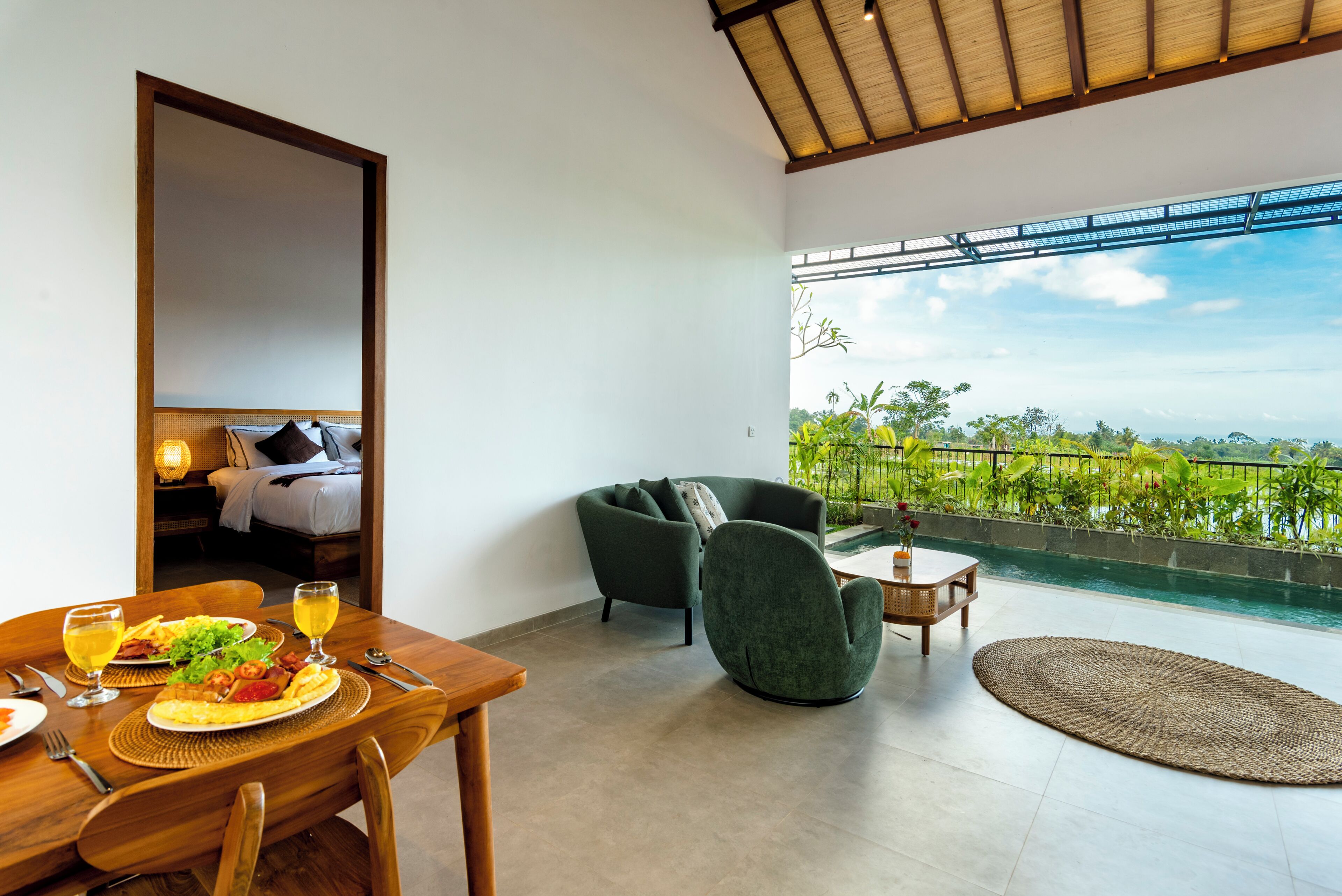 A room with a dining table set for a meal, a sofa, and a view of a pool and greenery.
