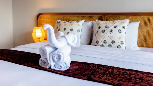 Een netjes opgemaakt bed met een decoratieve handdoek in de vorm van een olifant, een bekledingspaneel met een patroon en een bedlampje.