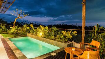 Una piscina con cubierta de madera y zona de estar, rodeada de abundante vegetación y vistas al cielo.