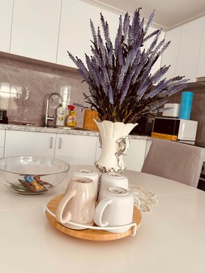 A kitchen with a white vase of purple flowers on a wooden tray, a glass bowl, and white ceramic cups. - New 2BR1B Flat+2 Free parking|Lake nearby&Hospital (North Parramatta)