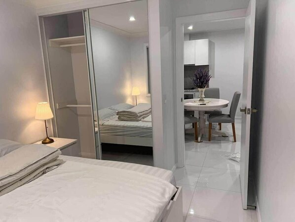 A hotel room with a bed, bedside table, lamp, and a view into the dining area.