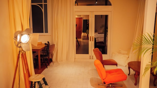 Ein Zimmer mit Esstisch, Stühlen, einer hellen Stehlampe, einem großen Fenster mit Vorhängen, einer Glastür, die in ein Schlafzimmer führt, und einer Zimmerpflanze.