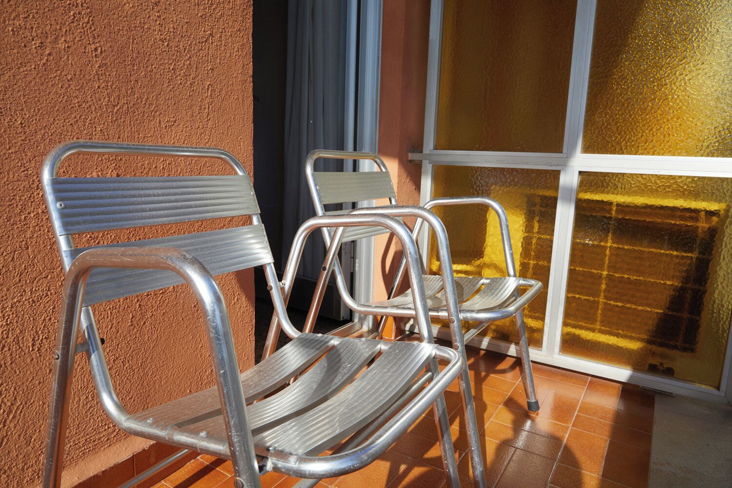 Two metal chairs with metal frames and wooden seats, positioned on a tiled floor near a window with a textured glass pane.