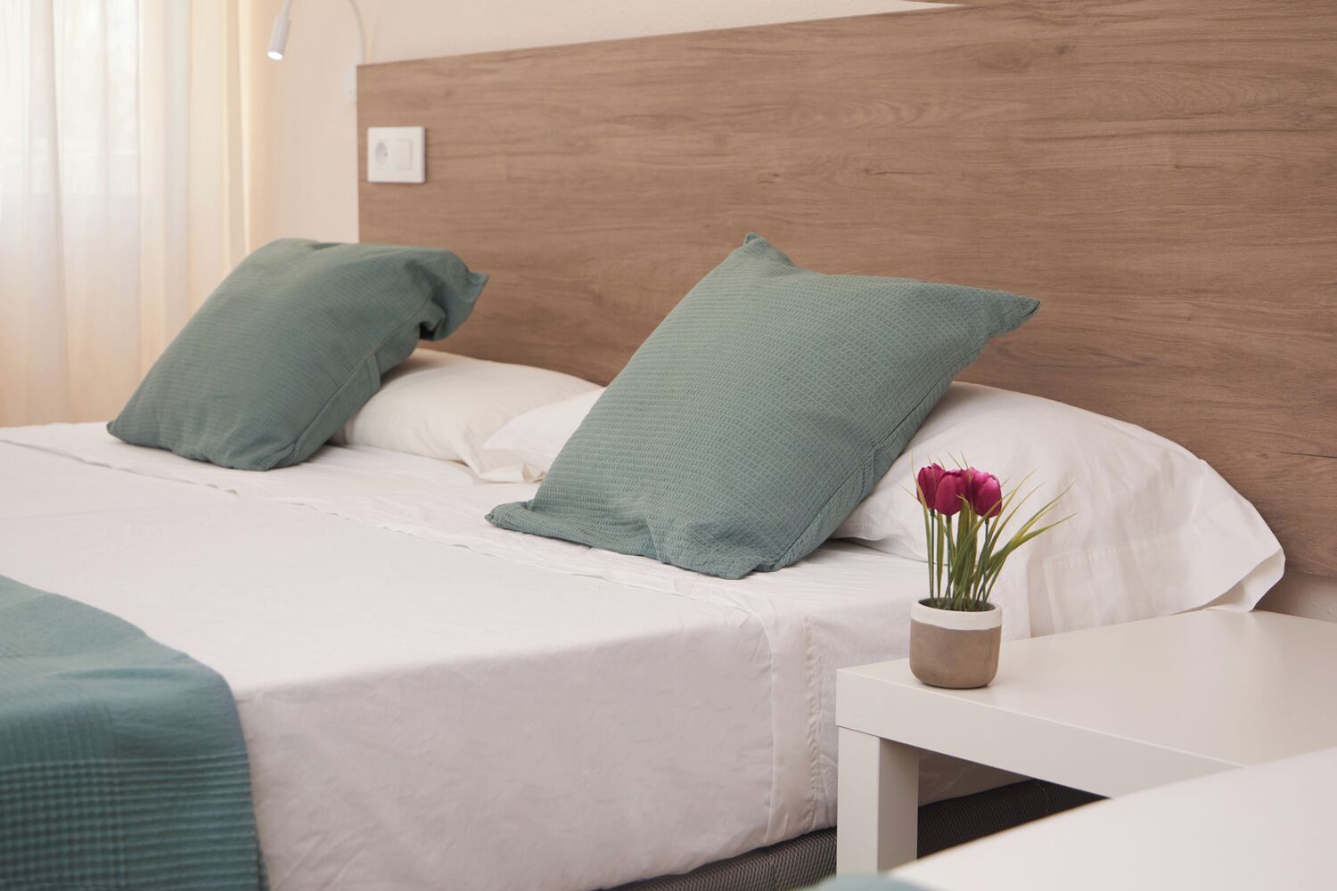 A neatly made bed with a white and green bedspread, a wooden headboard, and a bedside table with a vase of flowers.