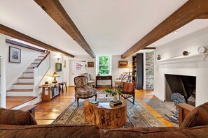 A spacious living room with a wooden staircase, a fireplace, and a view of greenery outside.