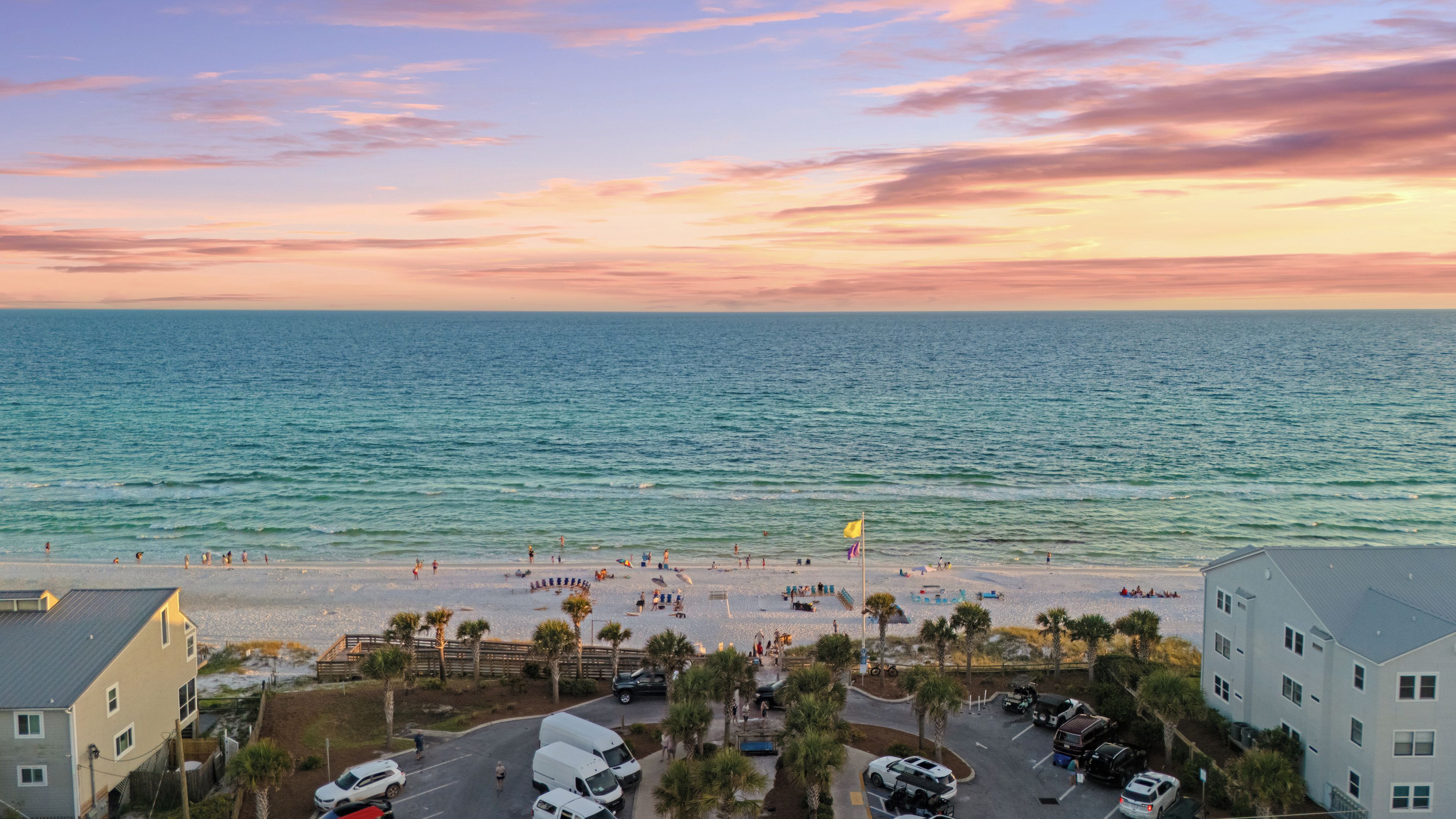 A beach view with a parking area, buildings, and a clear sky.