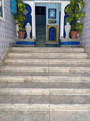 Marmeren trappen die leiden naar een blauwe deur met een plaquette, omlijst door potplanten en decoratieve tegels.