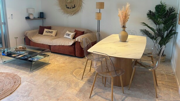 Una sala de estar con un sofá, una mesa de centro de vidrio, una mesa de comedor con sillas, una planta en maceta y una lámpara fijada a la pared.