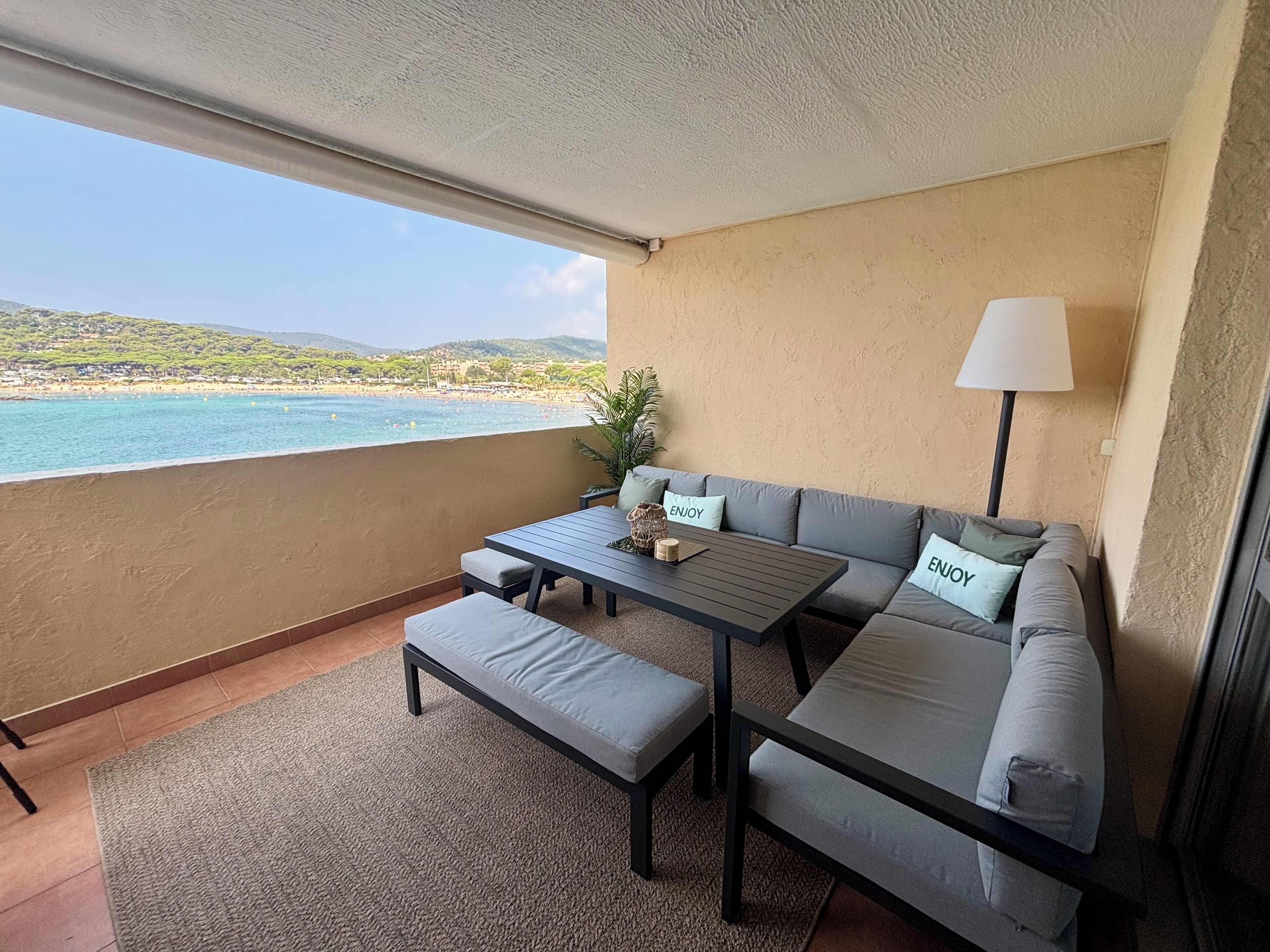 Un balcón con sofá, mesa y banco, con vistas a la playa y colinas.