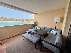 A balcony with a sofa, table, and bench, overlooking a beach and hills. - Spacious, luxury 3-bedroom apartment with ocean views and a pool (Bormes-les-Mimosas)