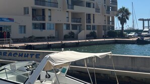 Un puerto deportivo con un barco blanco amarrado en primer plano, un edificio de varios pisos con balcones al fondo y un cielo azul despejado.