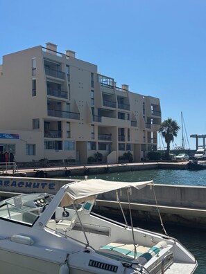 Ein Yachthafen mit einem weißen Boot im Vordergrund, ein mehrstöckiges Gebäude mit Balkonen im Hintergrund und einem strahlend blauen Himmel.