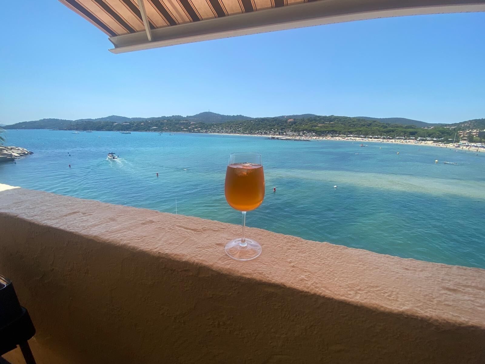 A glass of amber liquid on a balcony overlooking a coastal area with clear blue water, boats, and a distant shoreline.
