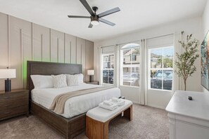 Ein modernes Schlafzimmer mit einem großen Bett, Nachttischen, einer Bank, einem Deckenventilator und Blick ins Freie durch große Fenster.