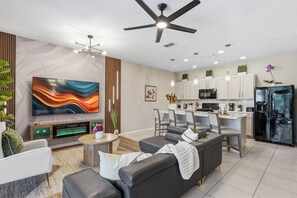 Ein modernes Wohnzimmer mit Flachbildfernseher, Kamin und Essbereich, im Hintergrund ist eine Küche zu sehen.