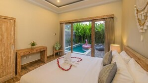 A hotel room with a bed, a wooden desk, a small table with a plant, a large window with a view of a swimming pool and greenery, and a decorative wall hanging. - Villa Dion Bali (Badung)