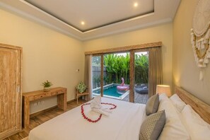 Ein Hotelzimmer mit einem Bett, einem Holztisch, einem kleinen Tisch mit einer Pflanze, einem großen Fenster mit Blick auf einen Swimmingpool und Grünflächen sowie einem Wandbild.