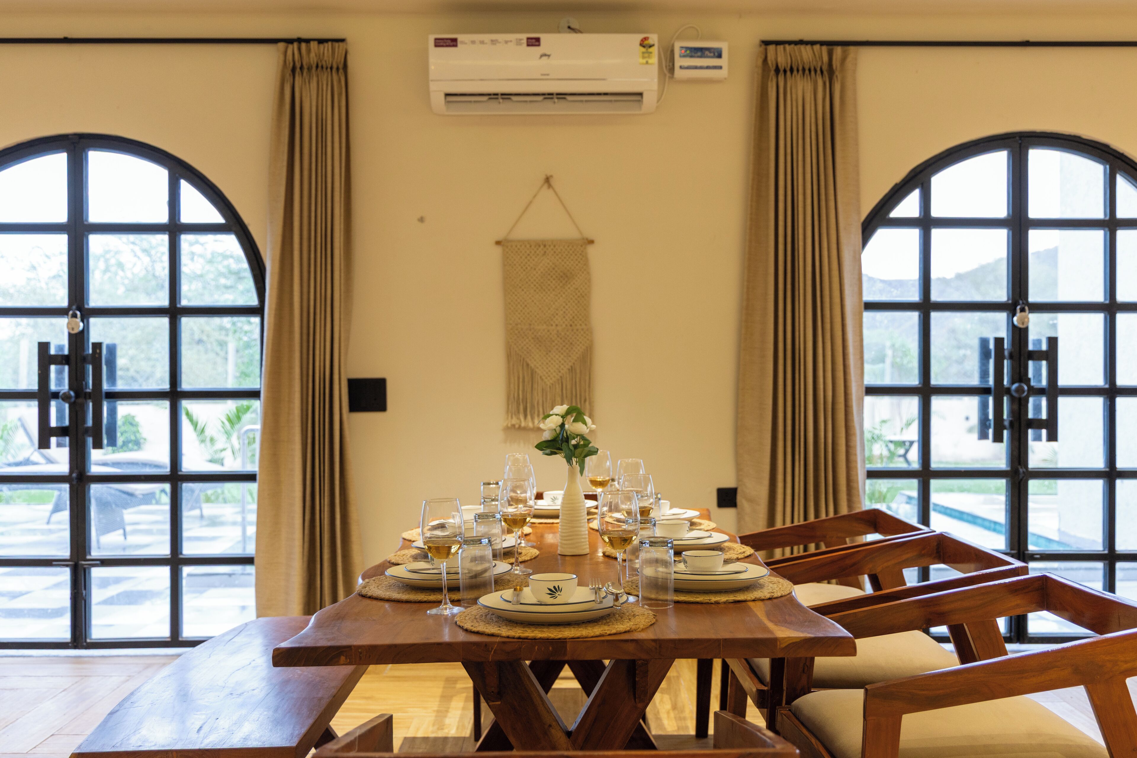 A dining table set for four with plates, glasses, and cutlery, flanked by two large windows with curtains.