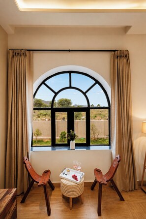 A room with a large arched window, two wooden chairs, a small table with a tray of fruit and glasses, and a potted plant.