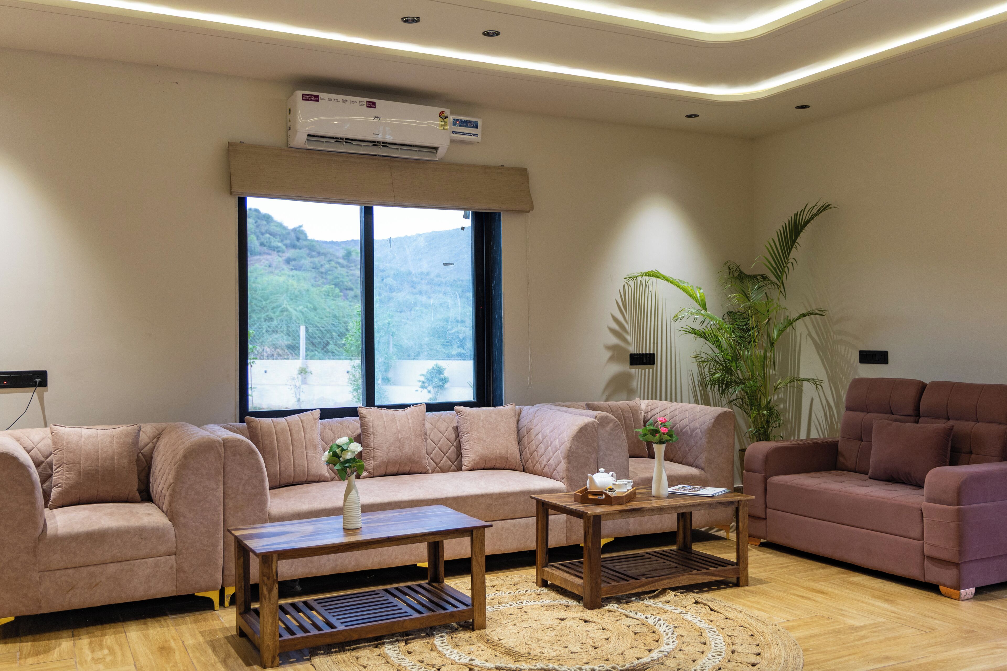 A living room with a beige sofa, two wooden coffee tables, a large window with a view of greenery, and an air conditioning unit on the ceiling.