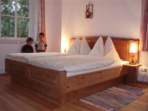 Ein hölzernes Bett mit weißen Leinentüchern in einem Zimmer mit Fenster, einem Nachttisch und einer Lampe.
