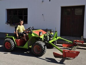 Ein Kind auf einem grünen Traktor mit roten Anbauten vor einem weißen Gebäude, das ein Fenster und eine Tür hat.