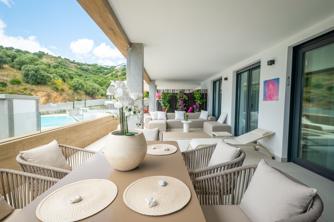 A balcony with wicker furniture, a large vase with flowers, and a view of the sea and hills.