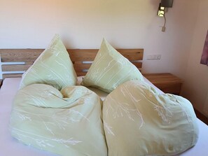 Ein Bett mit hölzernem Kopfteil, zwei Kissen mit Blättermotiv, ein Nachttisch und eine Wandleuchte.
