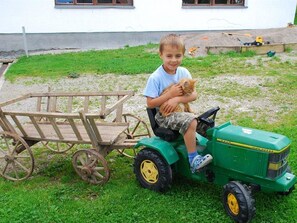 Ein Kind auf einem grünen Spielzeug-John-Deere-Traktor mit einem angehängten Holzwagen, draußen.