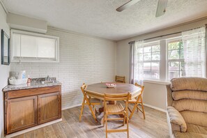 Ein Essbereich mit Holztisch und Stühlen, eine Küche mit weiß gefliester Spritzschutzwand und ein Fenster mit Vorhängen.
