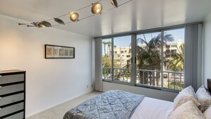 Ein Schlafzimmer mit großem Fenster, einem Bett mit floraler Bettwäsche, einer Kommode und einem gerahmten Bild an der Wand.
